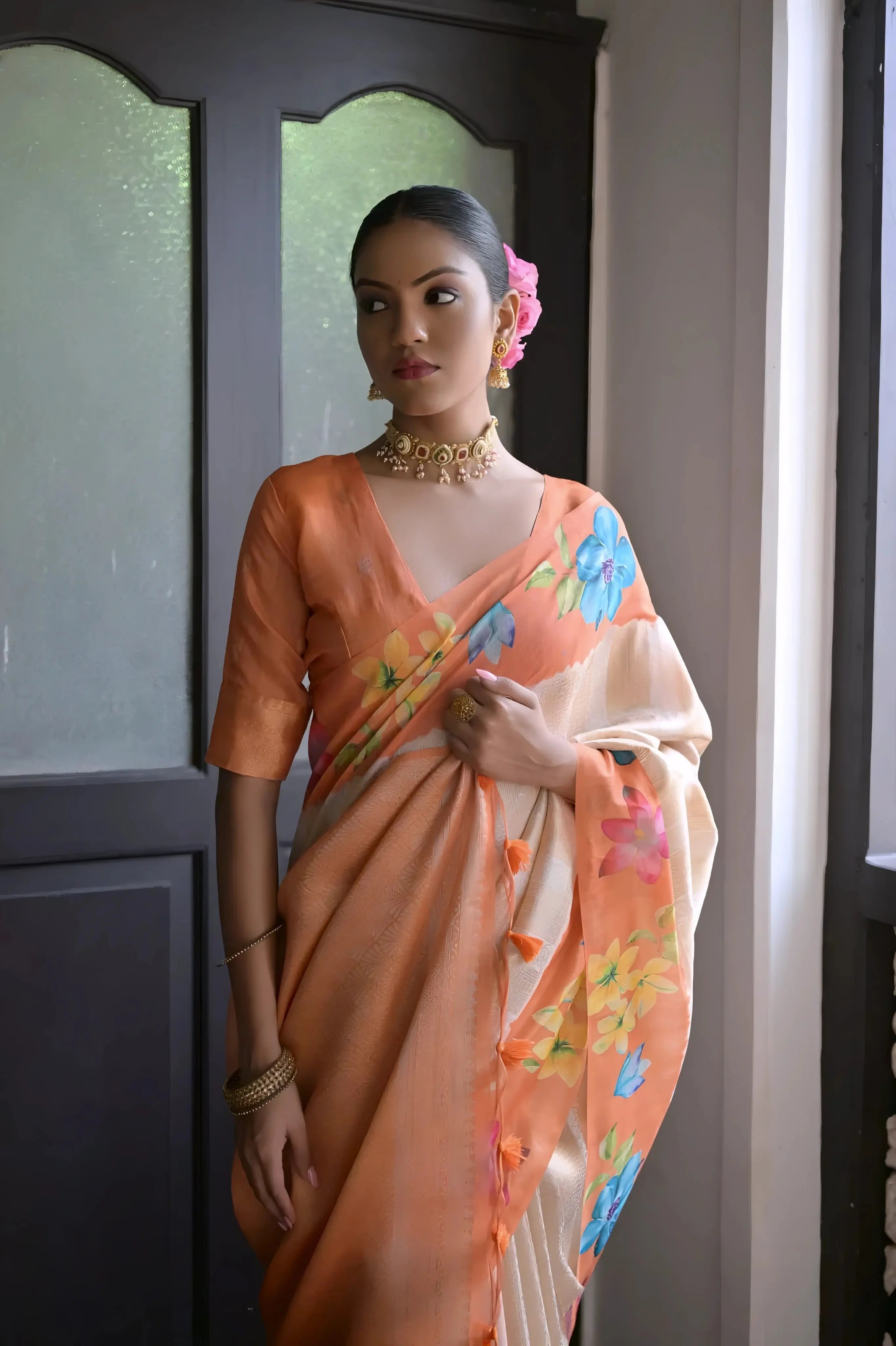A woman models a graceful Soft Silk Saree. The saree body is pale beige/cream with subtle Zari weaving, and features a peach/coral border. The shoulder and pallu area are decorated with delicate printed floral motifs in pink and green. She pairs it with a peach-colored blouse and a gold and pearl choker necklace.